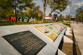 A series of informational plaques are arranged on a slanted concrete structure in a park setting. The backdrop features trees, open grassy areas, and scattered people, with large, colorful letters in the foliage spelling out 'CDMX'. A blue sky with clouds can be seen above the scene.
