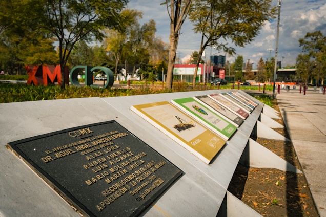 A series of informational plaques are arranged on a slanted concrete structure in a park setting. The backdrop features trees, open grassy areas, and scattered people, with large, colorful letters in the foliage spelling out 'CDMX'. A blue sky with clouds can be seen above the scene.