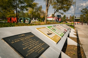 A series of informational plaques are arranged on a slanted concrete structure in a park setting. The backdrop features trees, open grassy areas, and scattered people, with large, colorful letters in the foliage spelling out 'CDMX'. A blue sky with clouds can be seen above the scene.