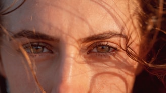 A close-up of a person's eyes and eyebrows, with sunlight casting shadows and highlighting the natural texture of the skin and hair.