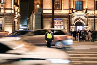A traffic officer stands in the middle of a busy intersection at night, wearing a reflective vest. Cars speed by, creating a motion blur effect. A group of people wait on the sidewalk near a brightly lit building.