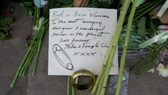 A handwritten note is placed among green flowers and stems, possibly as part of a memorial. The note, written with black ink, contains a message of condolence and appreciation, addressed to someone named Vivienne. Beside the note, there is a large gold ring and scattered multicolored beads. The scene conveys a sense of tribute and remembrance.