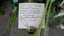 A handwritten note is placed among green flowers and stems, possibly as part of a memorial. The note, written with black ink, contains a message of condolence and appreciation, addressed to someone named Vivienne. Beside the note, there is a large gold ring and scattered multicolored beads. The scene conveys a sense of tribute and remembrance.