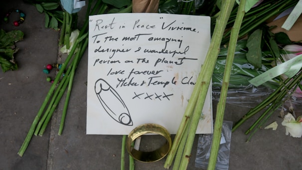 A handwritten note is placed among green flowers and stems, possibly as part of a memorial. The note, written with black ink, contains a message of condolence and appreciation, addressed to someone named Vivienne. Beside the note, there is a large gold ring and scattered multicolored beads. The scene conveys a sense of tribute and remembrance.