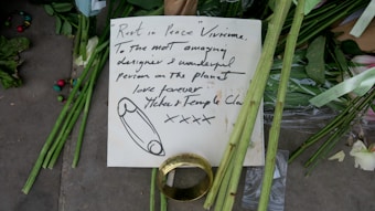 A handwritten note is placed among green flowers and stems, possibly as part of a memorial. The note, written with black ink, contains a message of condolence and appreciation, addressed to someone named Vivienne. Beside the note, there is a large gold ring and scattered multicolored beads. The scene conveys a sense of tribute and remembrance.