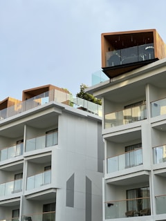 Modern multi-story building with a minimalist design featuring large glass balconies. The structure has unique wooden elements on the upper floors and is surrounded by clear sky. Lush greenery is visible on some of the balconies.