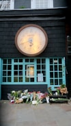 A large clock with the words 'world's end' is mounted above a building with turquoise window frames. Below, numerous bouquets of flowers are placed on the ground, suggesting a memorial or tribute. The background includes a dark slate wall with an upstairs window featuring small plants.