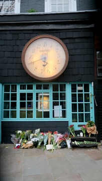 A large clock with the words 'world's end' is mounted above a building with turquoise window frames. Below, numerous bouquets of flowers are placed on the ground, suggesting a memorial or tribute. The background includes a dark slate wall with an upstairs window featuring small plants.