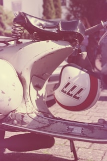 A close-up view of a vintage motorcycle with a weathered seat and a white helmet decorated with red and black graphics hanging from the handlebar. The scene is outdoors, with some trees and a partial view of a building in the blurred background, giving a retro aesthetic.