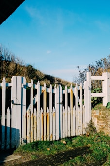 A worn white picket fence with noticeable rust stains stands under a vibrant blue sky. There is grassy terrain surrounding the fence, with some scattered dry leaves and sparse vegetation. The setting appears natural and slightly overgrown, with a rustic and aged appearance.