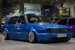 A customized blue Volkswagen GTI car with lowered suspension and shiny chrome wheels is parked indoors. The background includes several other cars, banners, and a sign with the text 'mobile.bg'. The area has an industrial or garage-like setting with overhead lighting.