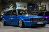 A customized blue Volkswagen GTI car with lowered suspension and shiny chrome wheels is parked indoors. The background includes several other cars, banners, and a sign with the text 'mobile.bg'. The area has an industrial or garage-like setting with overhead lighting.