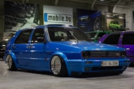 A customized blue Volkswagen GTI car with lowered suspension and shiny chrome wheels is parked indoors. The background includes several other cars, banners, and a sign with the text 'mobile.bg'. The area has an industrial or garage-like setting with overhead lighting.