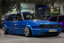 A customized blue Volkswagen GTI car with lowered suspension and shiny chrome wheels is parked indoors. The background includes several other cars, banners, and a sign with the text 'mobile.bg'. The area has an industrial or garage-like setting with overhead lighting.