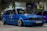A customized blue Volkswagen GTI car with lowered suspension and shiny chrome wheels is parked indoors. The background includes several other cars, banners, and a sign with the text 'mobile.bg'. The area has an industrial or garage-like setting with overhead lighting.