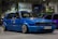 A customized blue Volkswagen GTI car with lowered suspension and shiny chrome wheels is parked indoors. The background includes several other cars, banners, and a sign with the text 'mobile.bg'. The area has an industrial or garage-like setting with overhead lighting.