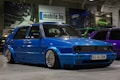 A customized blue Volkswagen GTI car with lowered suspension and shiny chrome wheels is parked indoors. The background includes several other cars, banners, and a sign with the text 'mobile.bg'. The area has an industrial or garage-like setting with overhead lighting.