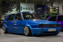 A customized blue Volkswagen GTI car with lowered suspension and shiny chrome wheels is parked indoors. The background includes several other cars, banners, and a sign with the text 'mobile.bg'. The area has an industrial or garage-like setting with overhead lighting.