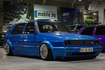 A customized blue Volkswagen GTI car with lowered suspension and shiny chrome wheels is parked indoors. The background includes several other cars, banners, and a sign with the text 'mobile.bg'. The area has an industrial or garage-like setting with overhead lighting.