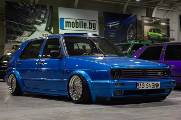 A customized blue Volkswagen GTI car with lowered suspension and shiny chrome wheels is parked indoors. The background includes several other cars, banners, and a sign with the text 'mobile.bg'. The area has an industrial or garage-like setting with overhead lighting.