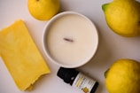 A creamy white candle set in a smooth round container is surrounded by fresh lemons and a bar of bright yellow soap. There is also a small bottle of lemon essential oil labeled 'PYRILA' positioned next to the candle.