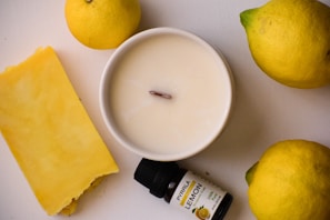 A creamy white candle set in a smooth round container is surrounded by fresh lemons and a bar of bright yellow soap. There is also a small bottle of lemon essential oil labeled 'PYRILA' positioned next to the candle.