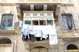 Several pieces of clothing hang out to dry on a clothesline outside a building. The building appears old, with weathered, stone walls and arched windows. An air conditioning unit is mounted on the wall next to the clothing. The area appears to be residential.