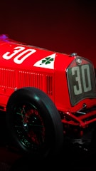 A vintage racing car with a prominent number 30 and a cloverleaf emblem on a vibrant red body. The visible section includes the front grille, a large spoked wheel, and part of the engine.