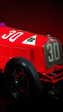 A vintage racing car with a prominent number 30 and a cloverleaf emblem on a vibrant red body. The visible section includes the front grille, a large spoked wheel, and part of the engine.