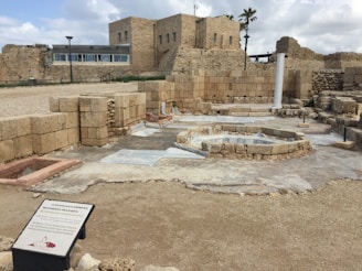 An ancient archaeological site featuring stone ruins and remnants of historical architecture. There are various levels of stonework with distinct rectangular blocks forming walls and pathways. A lone white column stands prominently amidst the ruins, surrounded by scattered stones and excavated sections. In the background, more intact buildings showcase the historical construction methods, set under a cloudy blue sky.