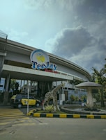 The entrance of a waterpark features a large sign with the name 'TeeJay Waterpark'. There is a sculpture of a giraffe near the entrance, surrounded by landscaping that includes a stone border and plants. A large, mushroom-shaped fountain is nearby. A taxi with advertising on its side is parked on the street, and several people can be seen walking near the entrance. The sky is overcast, with a mix of clouds and some light blue.