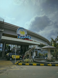 The entrance of a waterpark features a large sign with the name 'TeeJay Waterpark'. There is a sculpture of a giraffe near the entrance, surrounded by landscaping that includes a stone border and plants. A large, mushroom-shaped fountain is nearby. A taxi with advertising on its side is parked on the street, and several people can be seen walking near the entrance. The sky is overcast, with a mix of clouds and some light blue.