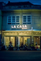 A cozy café named 'La Casa' is adorned with warm string lights and features a facade with a vintage architectural style. The café entrance has a couple of patrons sitting with their scooters parked in front of the establishment. The atmosphere inside seems inviting with framed artwork visible through the windows.