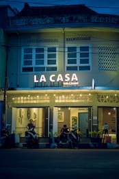 A cozy caf&eacute; named 'La Casa' is adorned with warm string lights and features a facade with a vintage architectural style. The caf&eacute; entrance has a couple of patrons sitting with their scooters parked in front of the establishment. The atmosphere inside seems inviting with framed artwork visible through the windows.