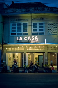 A cozy café named 'La Casa' is adorned with warm string lights and features a facade with a vintage architectural style. The café entrance has a couple of patrons sitting with their scooters parked in front of the establishment. The atmosphere inside seems inviting with framed artwork visible through the windows.