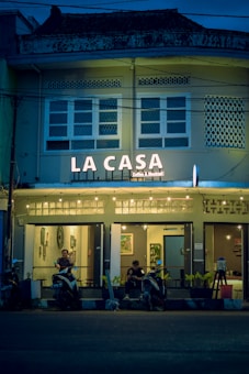 A cozy café named 'La Casa' is adorned with warm string lights and features a facade with a vintage architectural style. The café entrance has a couple of patrons sitting with their scooters parked in front of the establishment. The atmosphere inside seems inviting with framed artwork visible through the windows.