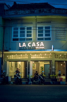 A cozy café named 'La Casa' is adorned with warm string lights and features a facade with a vintage architectural style. The café entrance has a couple of patrons sitting with their scooters parked in front of the establishment. The atmosphere inside seems inviting with framed artwork visible through the windows.