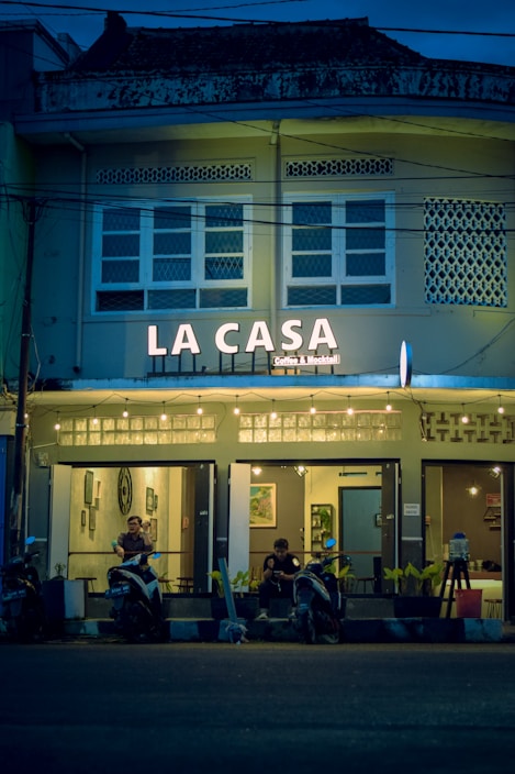 A cozy caf&eacute; named 'La Casa' is adorned with warm string lights and features a facade with a vintage architectural style. The caf&eacute; entrance has a couple of patrons sitting with their scooters parked in front of the establishment. The atmosphere inside seems inviting with framed artwork visible through the windows.