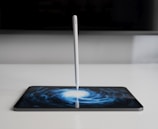 A stylus stands vertically on a tablet screen, which displays an artistic swirling pattern resembling a galaxy. The setting is minimalistic with muted colors, focused on the contrast between the white stylus and the dark screen art.