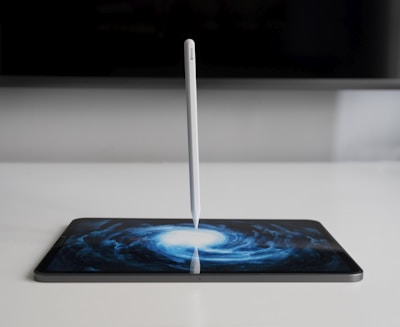 A stylus stands vertically on a tablet screen, which displays an artistic swirling pattern resembling a galaxy. The setting is minimalistic with muted colors, focused on the contrast between the white stylus and the dark screen art.