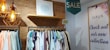 A clothing store display features a wooden wall with a drawing framed above. Hanging clothes in various pastel and neutral colors are visible on a rack. A 50% off sign is placed above the rack. Warm industrial-style pendant lights hang from the ceiling. On the right, a poster on the wall advertises a new collection with a large 'SALE' sign above it.