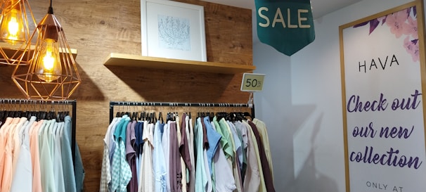 A clothing store display features a wooden wall with a drawing framed above. Hanging clothes in various pastel and neutral colors are visible on a rack. A 50% off sign is placed above the rack. Warm industrial-style pendant lights hang from the ceiling. On the right, a poster on the wall advertises a new collection with a large 'SALE' sign above it.