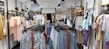 A clothing store with racks of various garments, including shirts, blouses, and pants on both sides of a central aisle. Sale signs are visible, with light tones of pastel colors and some printed patterns on the clothing.