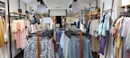 A clothing store with racks of various garments, including shirts, blouses, and pants on both sides of a central aisle. Sale signs are visible, with light tones of pastel colors and some printed patterns on the clothing.
