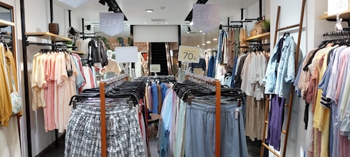 A clothing store with racks of various garments, including shirts, blouses, and pants on both sides of a central aisle. Sale signs are visible, with light tones of pastel colors and some printed patterns on the clothing.
