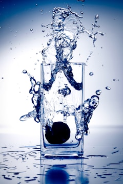 A dynamic splash of water erupts from a tall, clear glass as a dark object plunges into the liquid. Water droplets scatter in different directions with a vivid, energetic motion, set against a blue-hued background.