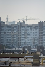 An urban landscape featuring a large residential building with multiple windows and balconies in the background. Several construction cranes rise into the sky, indicating ongoing development or construction. The foreground shows a rooftop with industrial equipment, including vents and ducts.