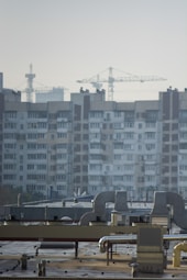 An urban landscape featuring a large residential building with multiple windows and balconies in the background. Several construction cranes rise into the sky, indicating ongoing development or construction. The foreground shows a rooftop with industrial equipment, including vents and ducts.