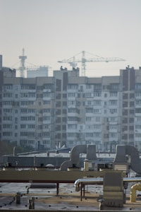 An urban landscape featuring a large residential building with multiple windows and balconies in the background. Several construction cranes rise into the sky, indicating ongoing development or construction. The foreground shows a rooftop with industrial equipment, including vents and ducts.