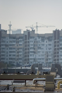 An urban landscape featuring a large residential building with multiple windows and balconies in the background. Several construction cranes rise into the sky, indicating ongoing development or construction. The foreground shows a rooftop with industrial equipment, including vents and ducts.